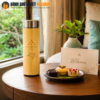 Bình giữ nhiệt vỏ tre đặt hài hòa trong không gian resort cạnh đĩa bánh ngọt và thiệp chào mừng.