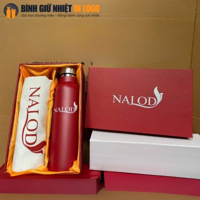 Cận cảnh bộ sản phẩm với bình giữ nhiệt đỏ mịn và túi canvas cuộn gọn trong hộp quà xi đỏ lót lụa vàng