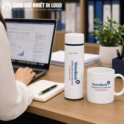Bình giữ nhiệt và cốc sứ in logo VietinBank đặt trên bàn làm việc gỗ bên cạnh laptop đang mở