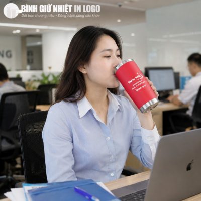 Nhân viên văn phòng nữ tập trung làm việc bên chiếc máy tính và thưởng thức đồ uống từ ly giữ nhiệt đỏ